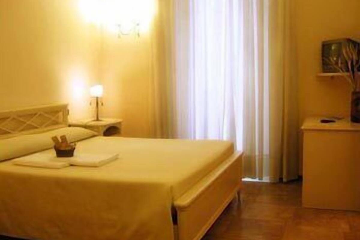 Ein Hotelzimmer mit einem Bett, einem Nachttisch mit Lampe, einem kleinen Tisch mit Vase und einem Fenster mit Vorhängen.