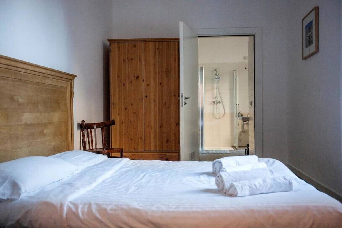 Une chambre avec une armoire en bois, une chaise, un lit avec du linge de lit blanc et une douche visible à travers une porte en verre.