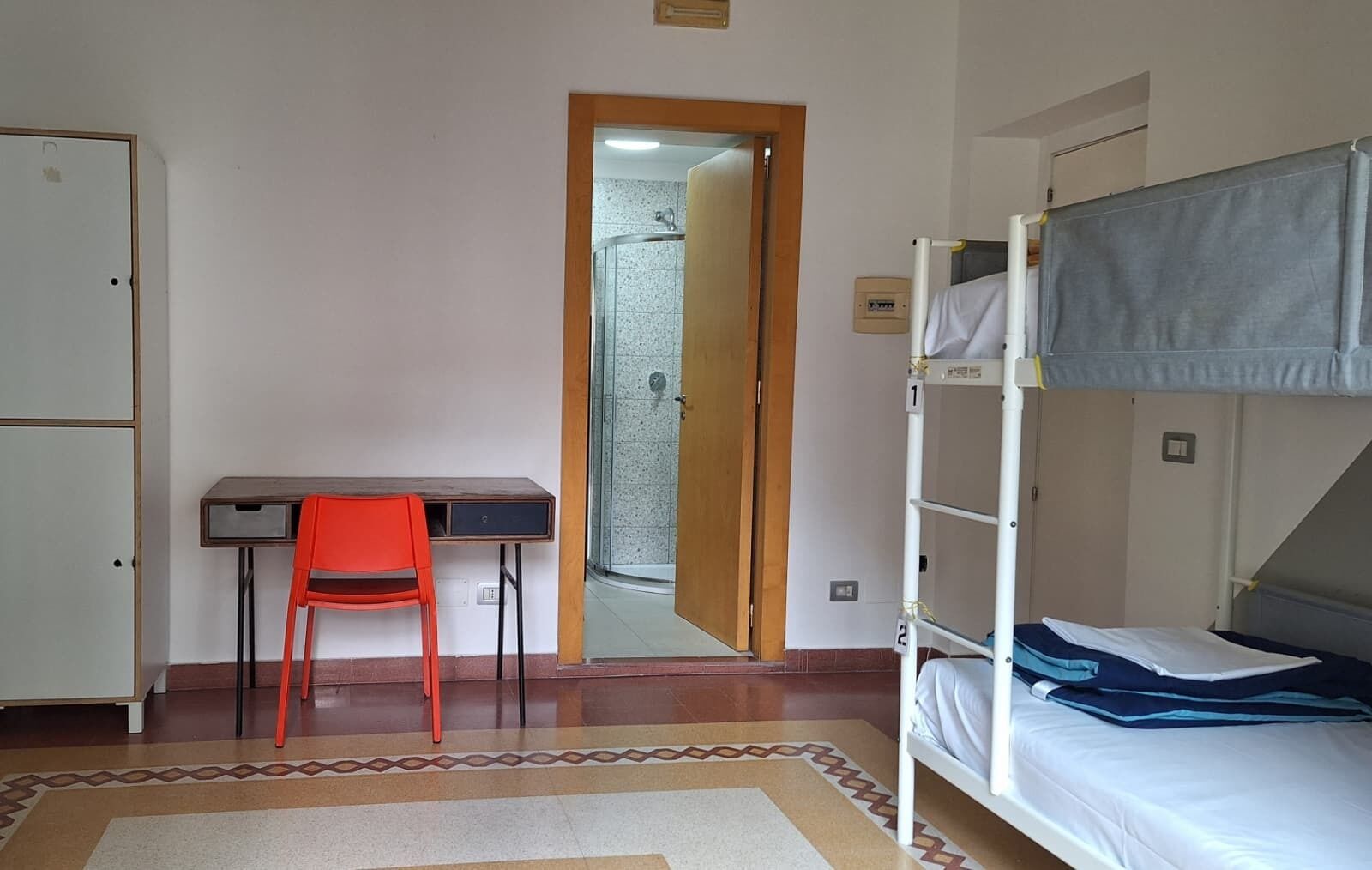 A room with a bunk bed, a desk with a chair, a wardrobe, and a shower area.