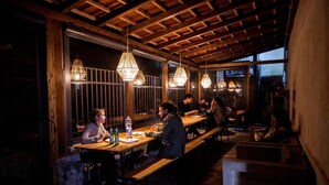 Een sfeervol verlicht restaurant met houten balken en hangende lampen. Tafels zijn gedekt met flessen en glazen, wat een informele eetomgeving suggereert.
