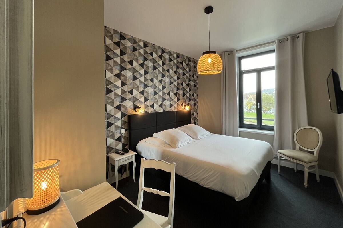 A hotel room with a bed, bedside tables, a chair, a desk, a lamp, and a window with a view.