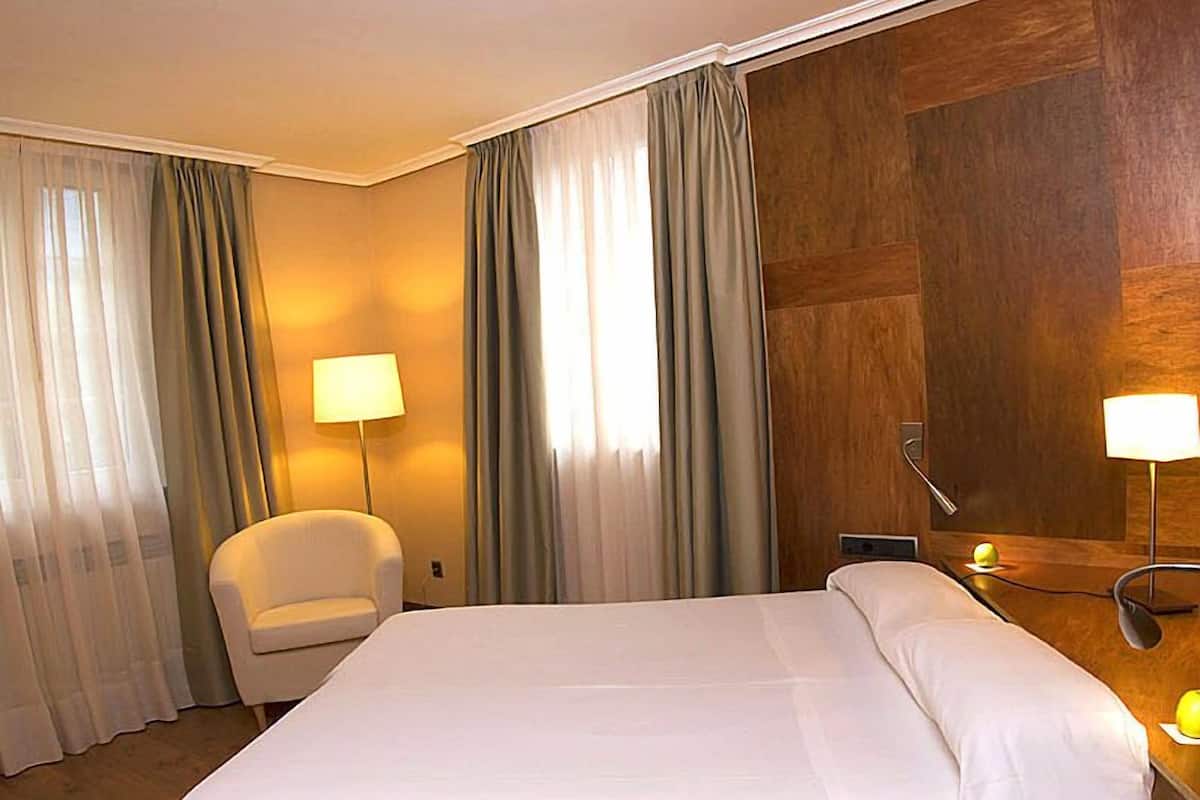 Una habitación de hotel con una cama grande, una silla, dos lámparas y un cabecero de madera.