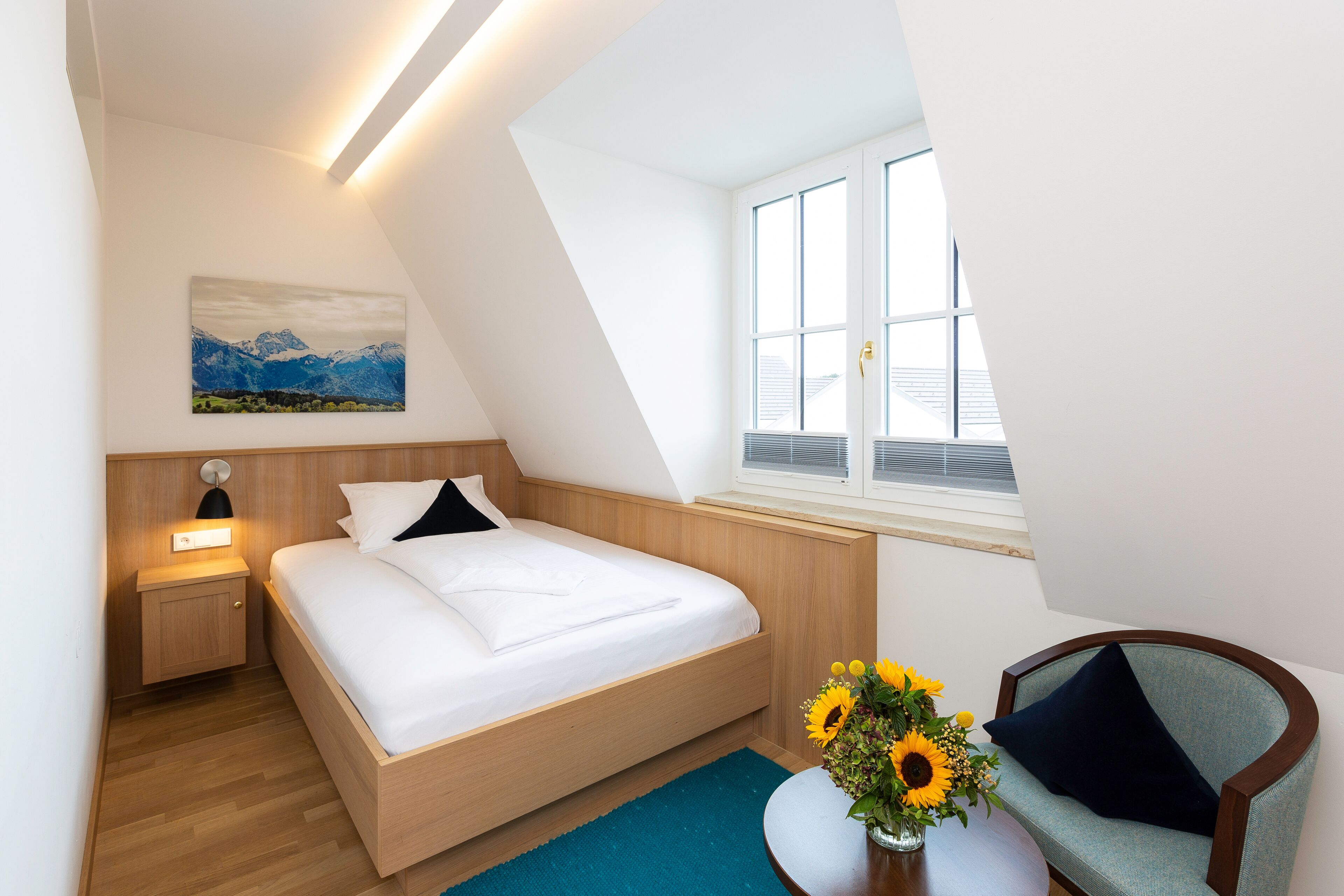 Ein modernes Schlafzimmer mit einem Holzbett, einem Nachttisch, einem Schreibtisch mit einer Vase voll Sonnenblumen und einem Fenster mit Bergblick.