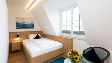 A modern bedroom with a wooden bed, a nightstand, a desk with a vase of sunflowers, and a window with a view of mountains.