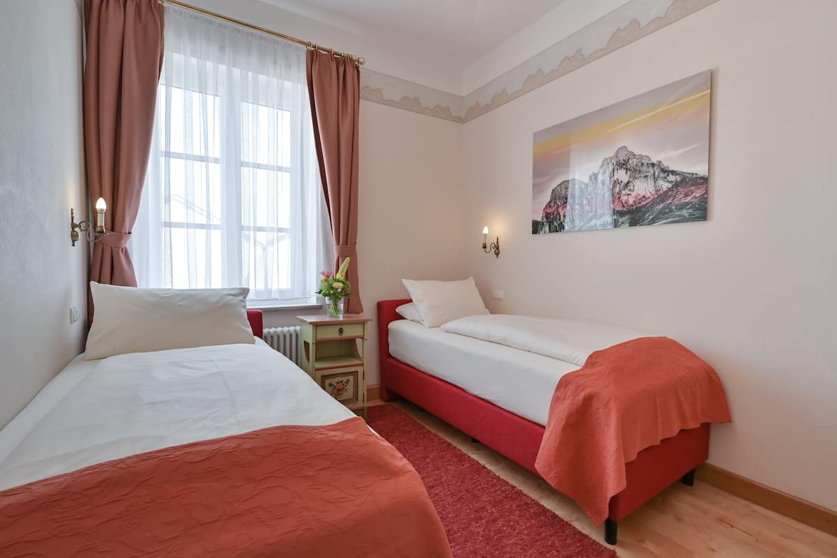Habitación de hotel con dos camas, una ventana con cortinas, un radiador, una mesita con un florero y un cuadro en la pared.