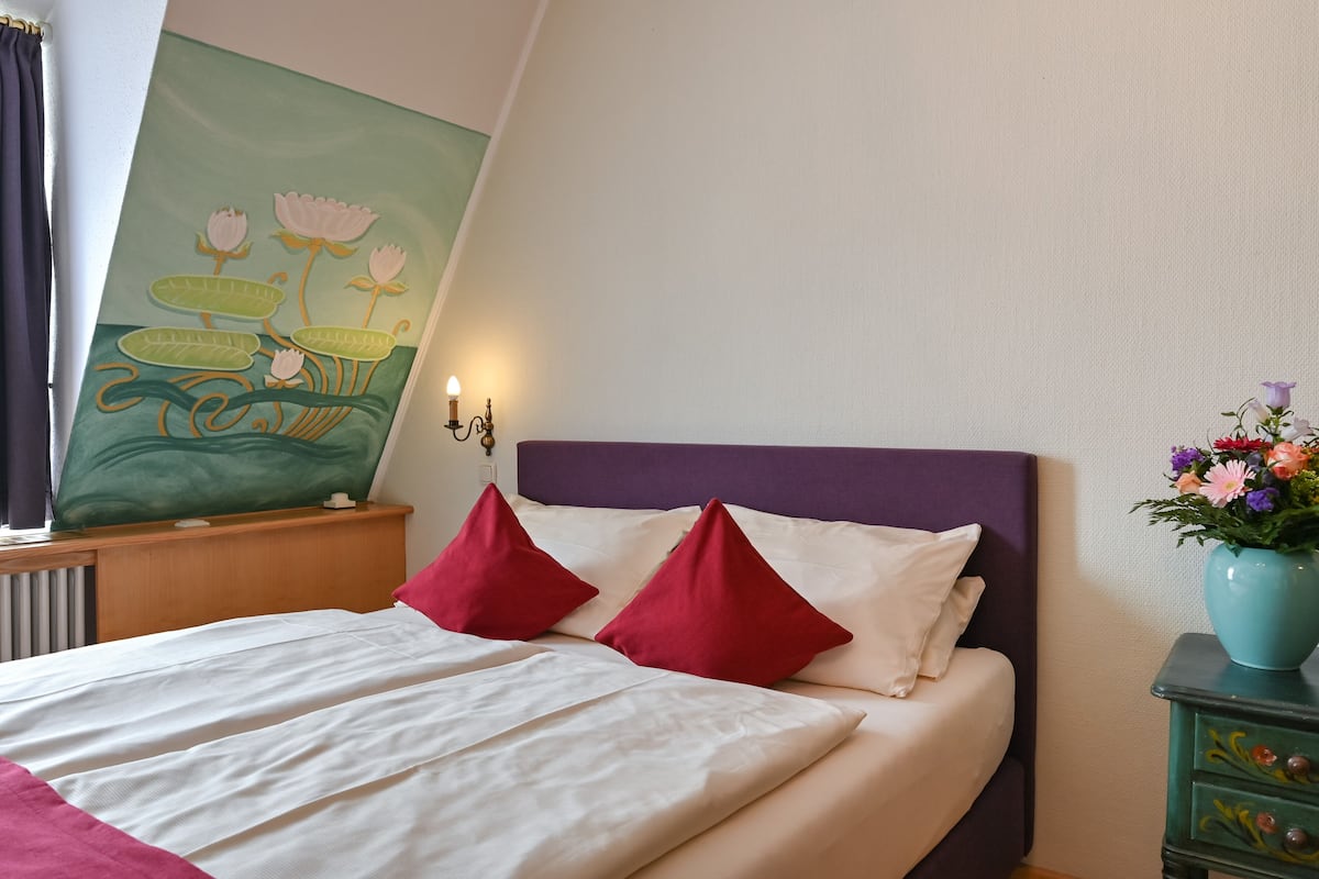 Una cama bien hecha con almohadas blancas y rojas, un cabecero morado y un cuadro de flores en la pared.