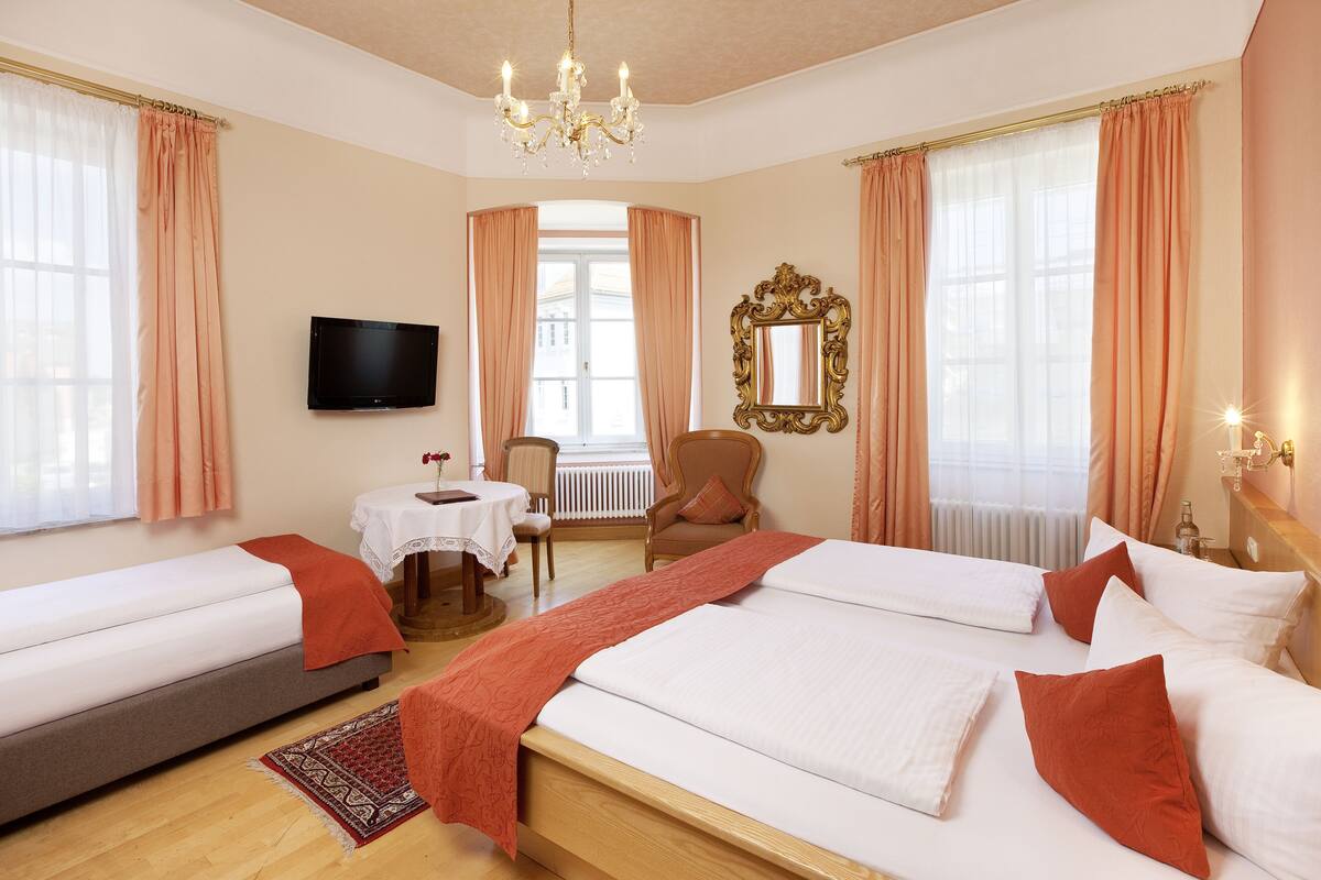 Habitación de hotel con una cama grande, una cama más pequeña, televisión, silla, mesa y espejo.