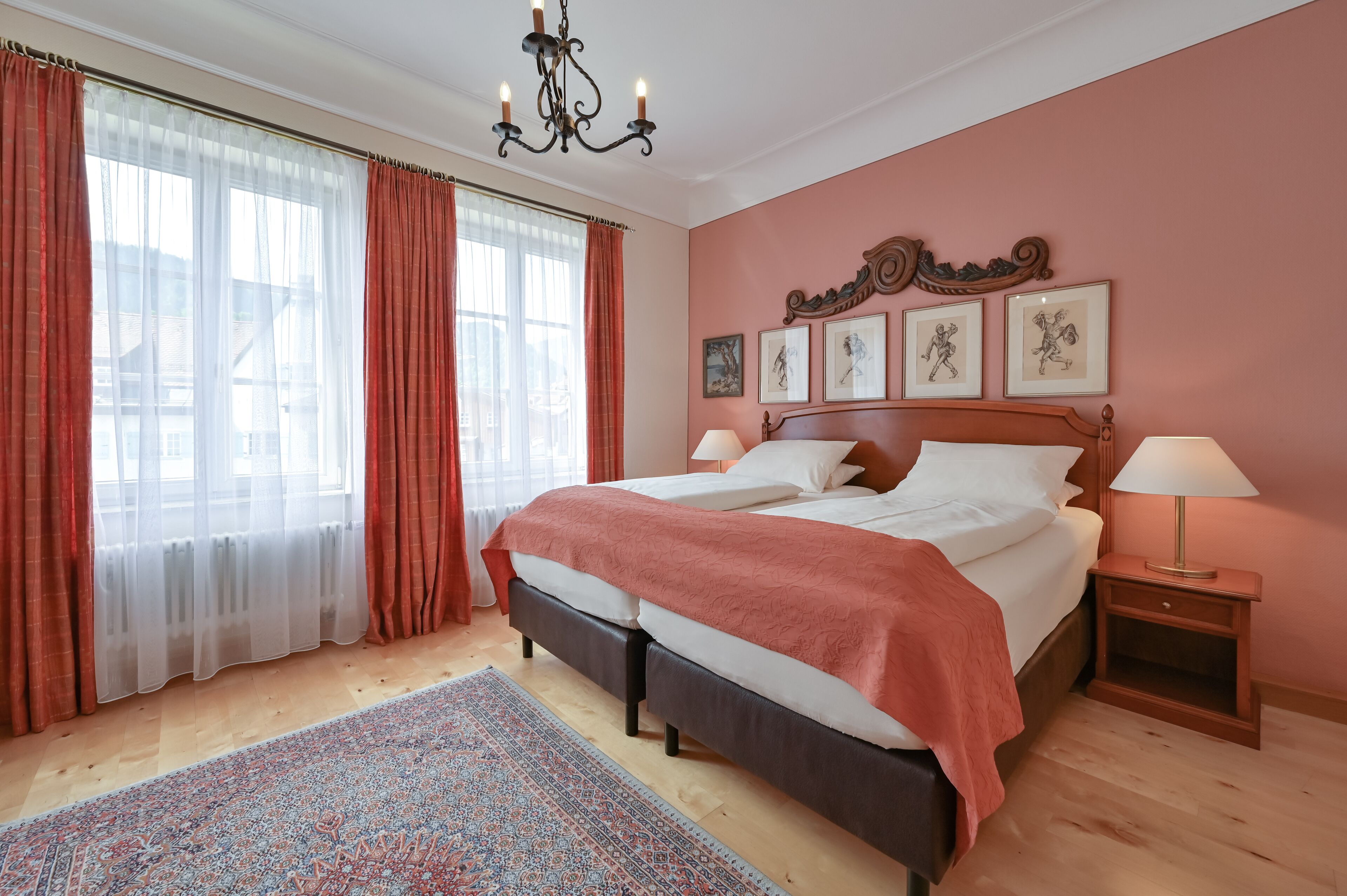 A bedroom with a large bed, two bedside tables, a chandelier, and a view of the outside through sheer curtains.