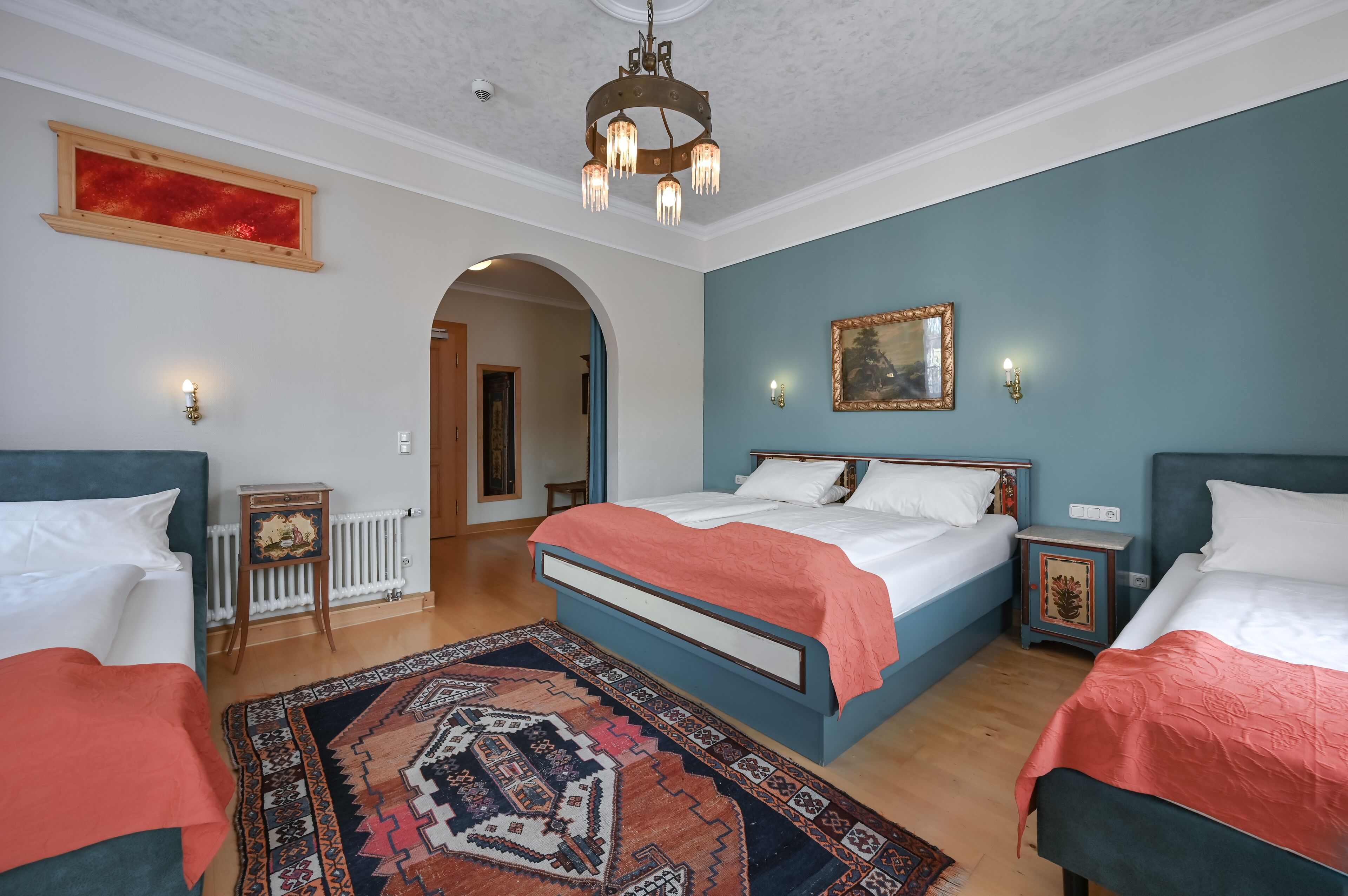 A bedroom with two beds, a chandelier, a patterned rug, and a painting on the wall.