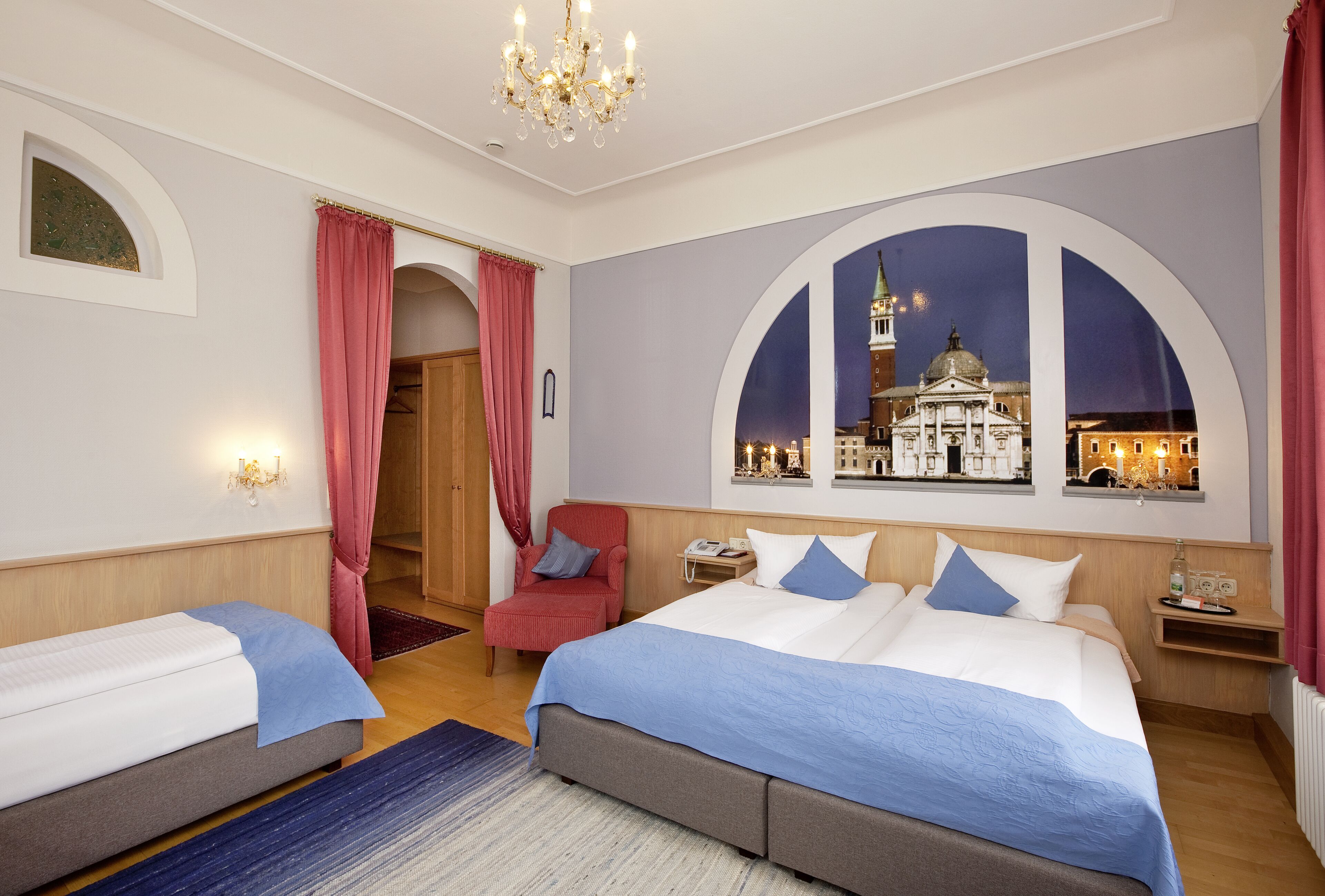 A hotel room with two beds, a view of a cityscape, a chandelier, and a red chair.