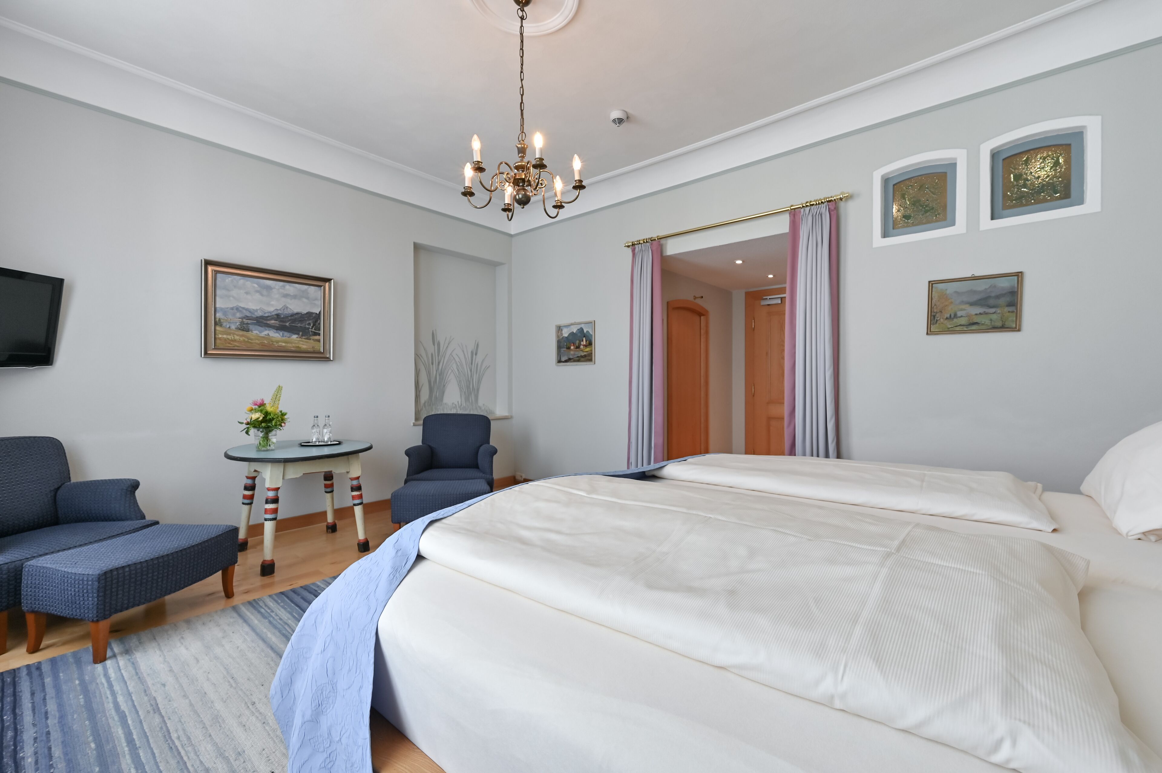 A bedroom with a large bed, a chair, a small table with a vase, and a TV on the wall.