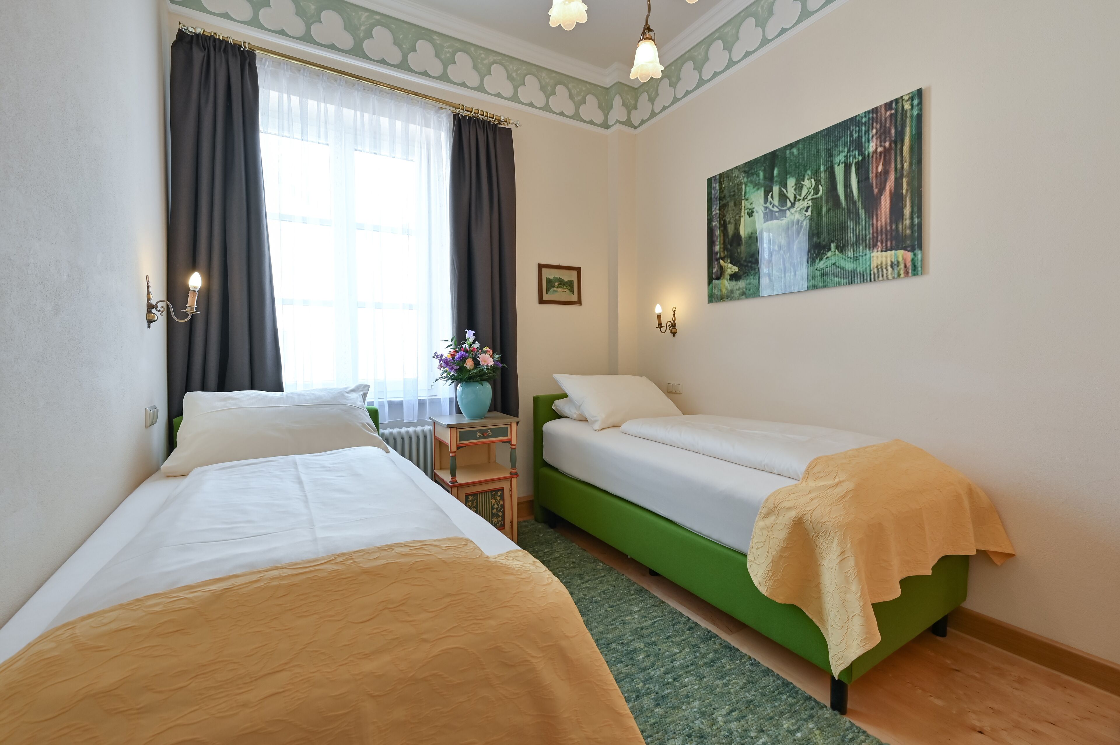 Habitación de hotel con dos camas individuales, una mesita con un jarrón de flores, un cuadro en la pared y una ventana con cortinas.