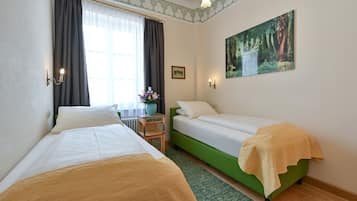 Habitación de hotel con dos camas individuales, una mesita con un jarrón de flores, un cuadro en la pared y una ventana con cortinas.