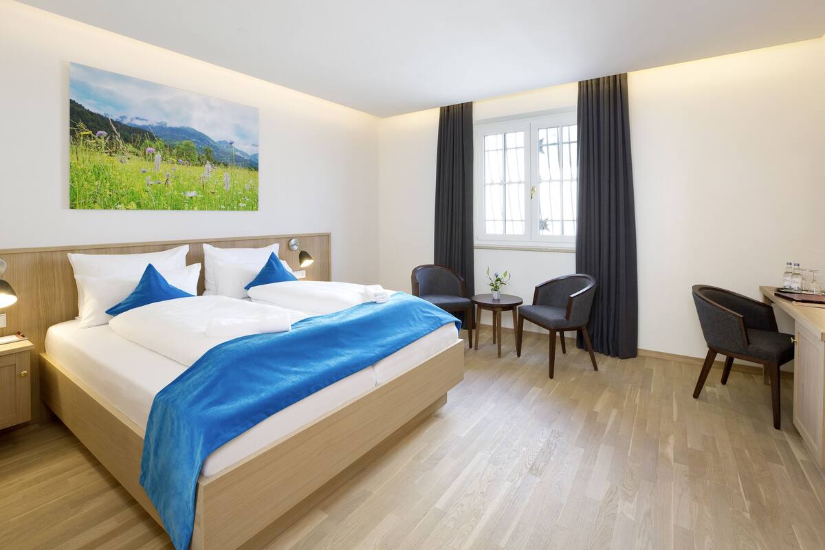 Habitación de hotel con una cama grande, un escritorio, dos sillas, una mesita y una ventana con cortinas.