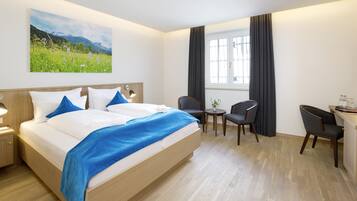 A hotel room with a large bed, a desk, two chairs, a small table, and a window with curtains.