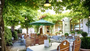 Een terras met houten tafels en stoelen, overdekt door een groene parasol, omringd door weelderig groen en grote ramen.