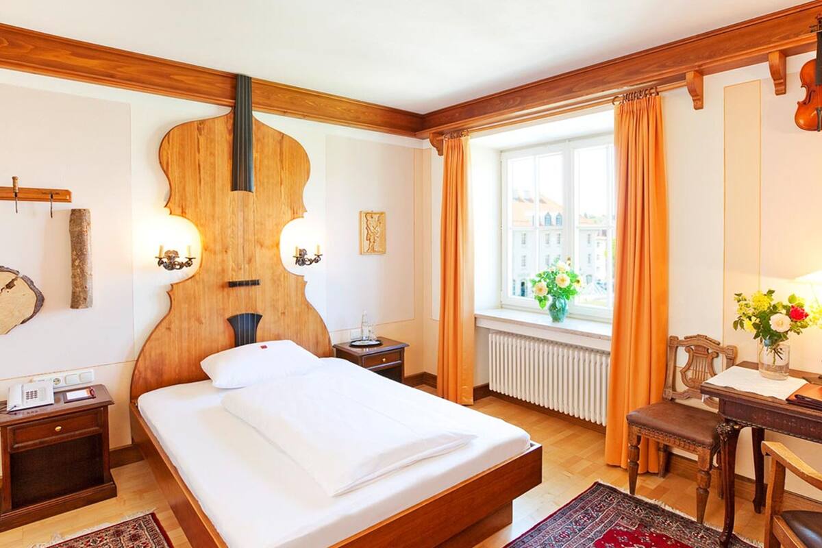 Habitación de hotel con una cama grande, cabecero de madera, una mesa pequeña con una silla, una ventana con cortinas y un violín colgado en la pared.