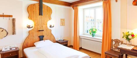 Habitación de hotel con una cama grande, cabecero de madera, una mesa pequeña con una silla, una ventana con cortinas y un violín colgado en la pared.