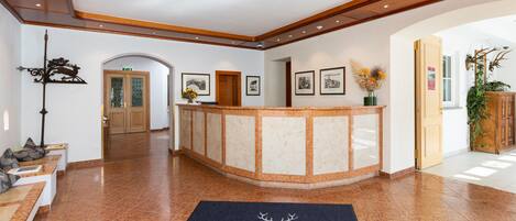 A hotel lobby with a reception desk, a blue carpet with the hotel name, a wooden reception desk, framed pictures on the wall, and a plant.