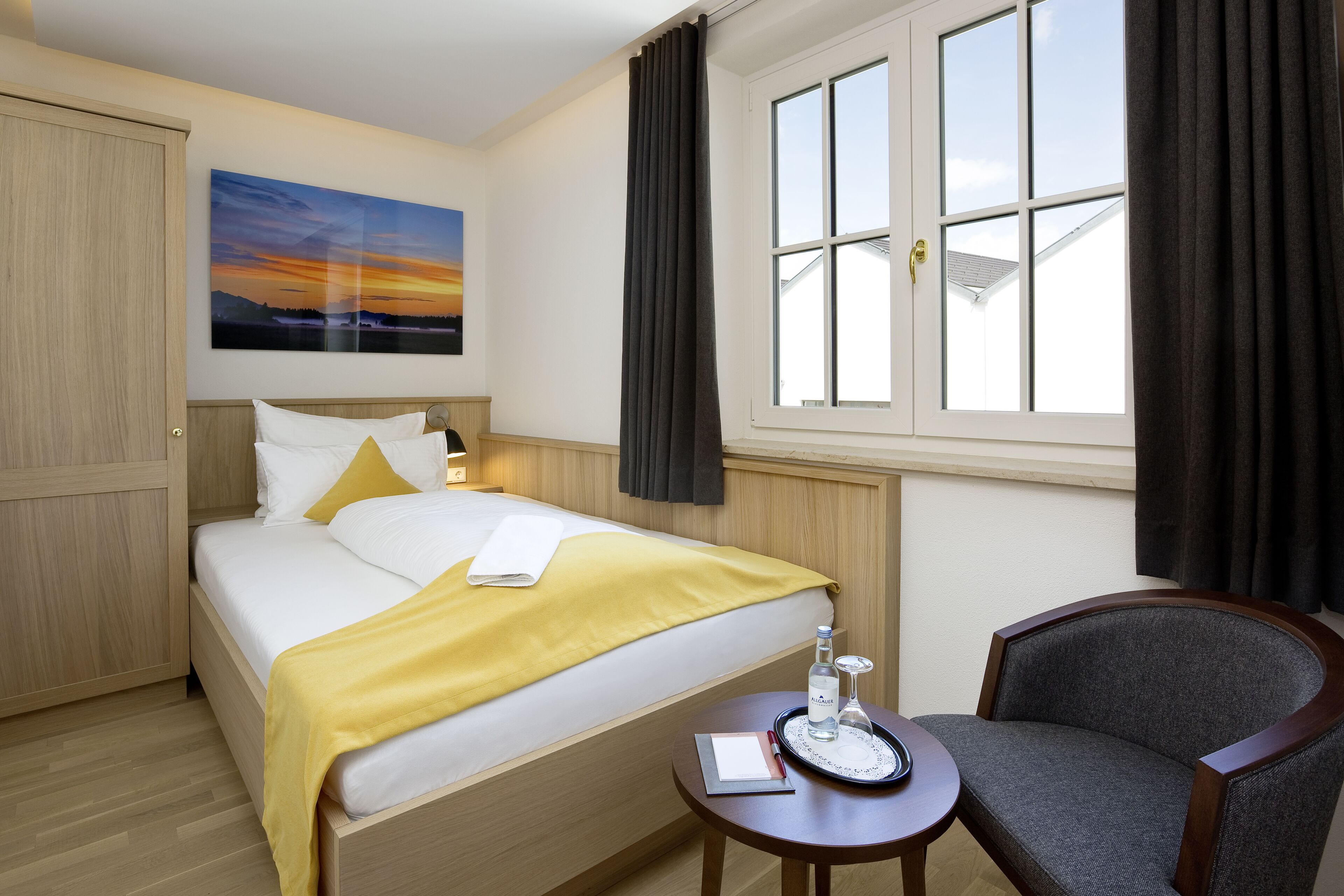 Een hotelkamer met een bed, een stoel, een klein tafeltje met een fles en glas, en een raam met gordijnen.