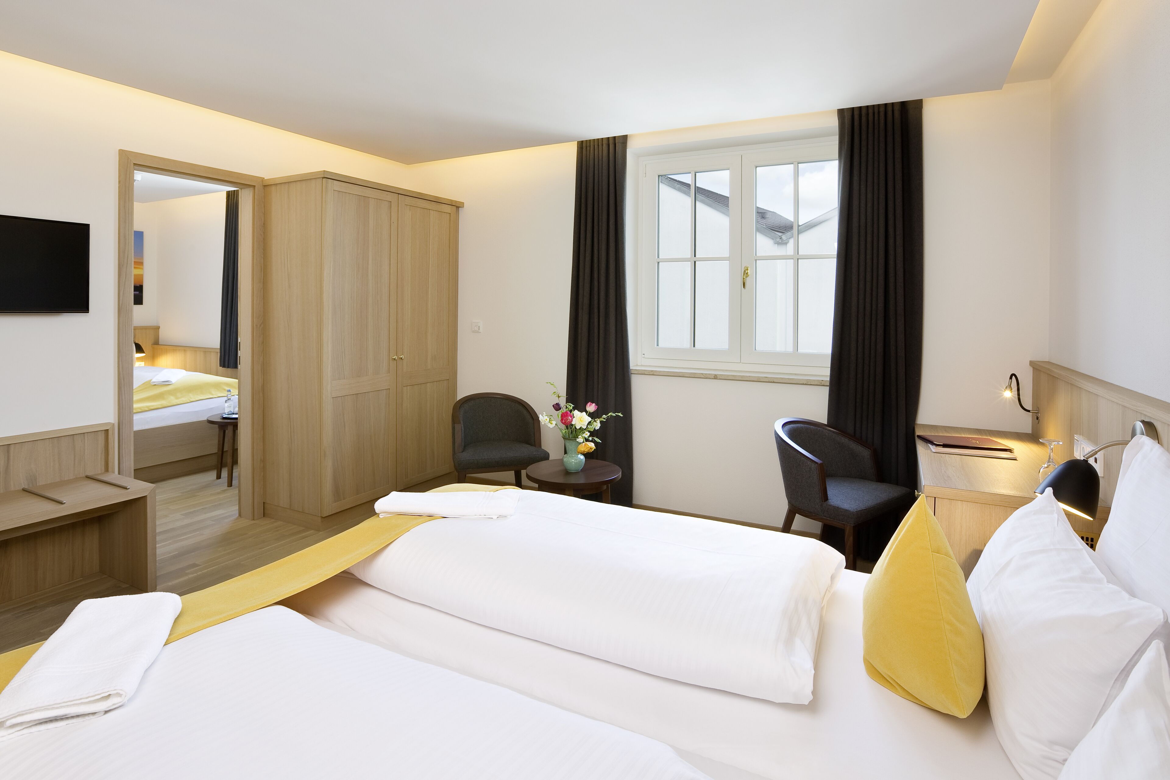A hotel room with two beds, a desk, a chair, a small table with a vase of flowers, and a window with curtains.
