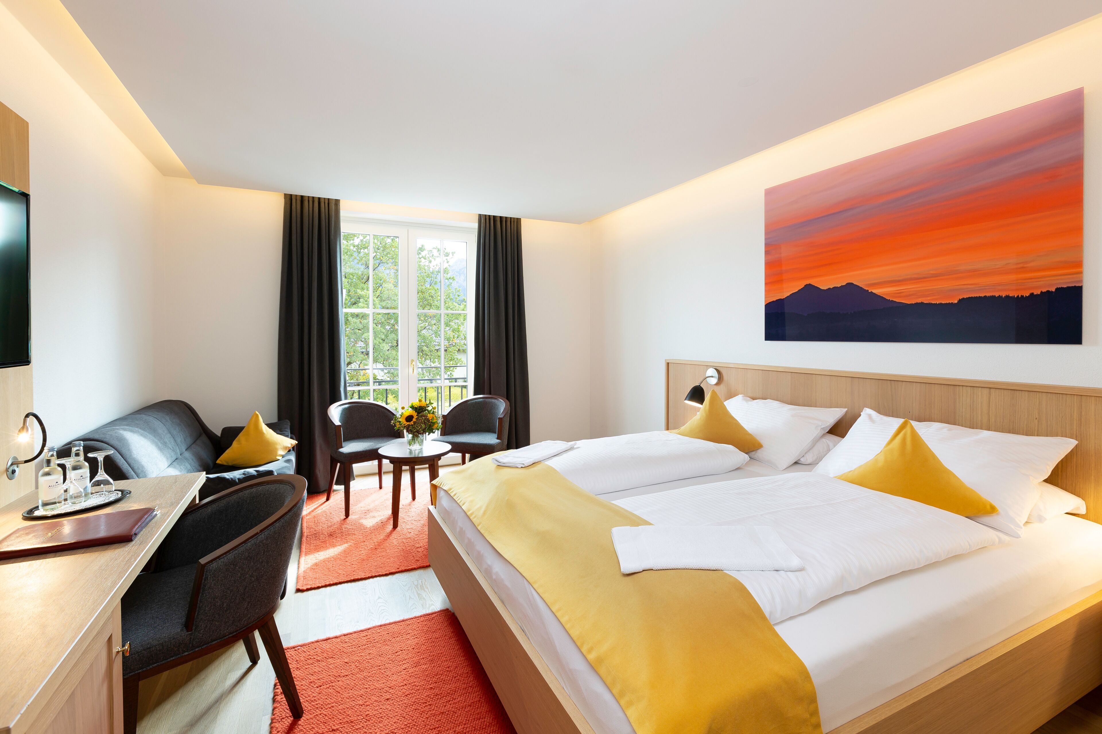 A hotel room with a bed, a desk, a chair, a small table with a vase of flowers, and a painting of a sunset on the wall.