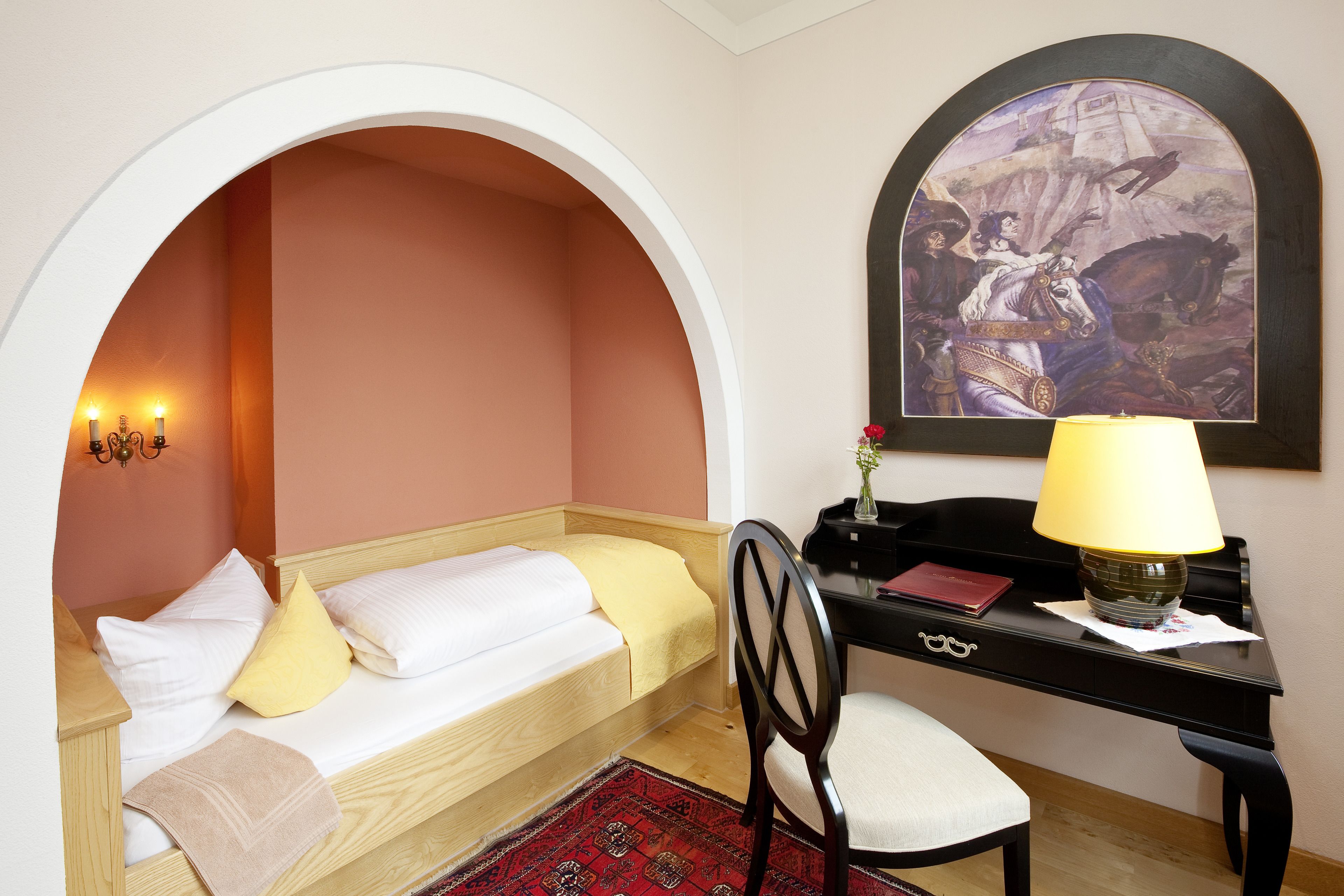 A bedroom with a single bed, a desk, a chair, a lamp, and a painting on the wall.