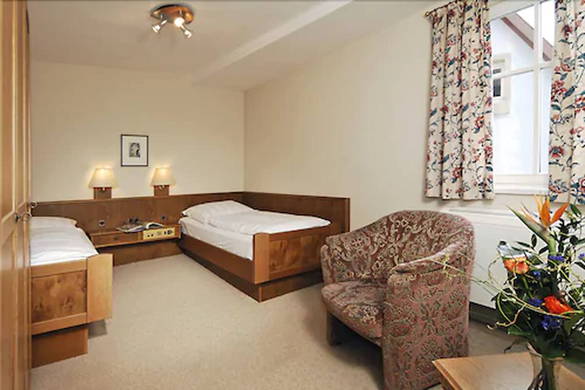 A hotel room with two single beds, a wooden nightstand with lamps, a patterned armchair, and a floral curtain.