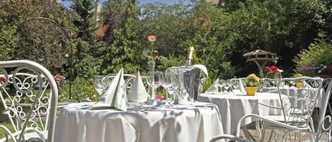 An outdoor dining area with round tables covered in white tablecloths, surrounded by white metal chairs.