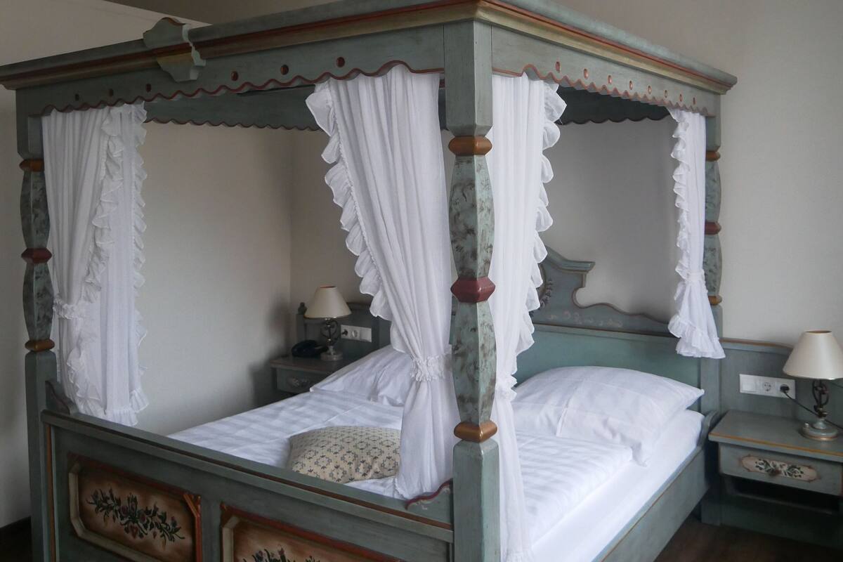 A four-poster canopy bed with white bedding, a floral headboard, and a nightstand with a lamp.