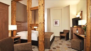 A hotel room with a large bed, two armchairs, a wooden accent wall, a small desk with a lamp, and a television.
