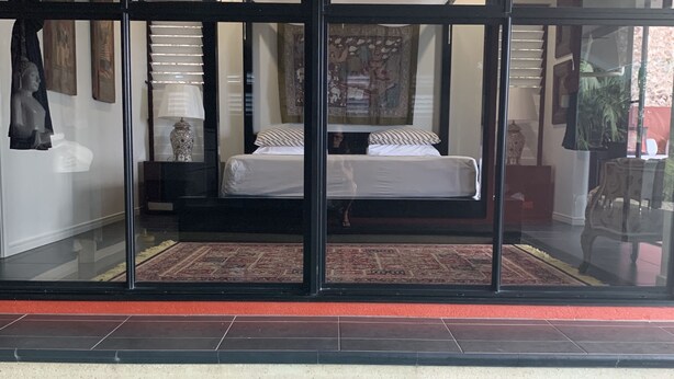 Quarto com divisória de vidro, uma cama, ventoinha no teto e vista para o exterior.