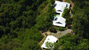 Vista aérea de uma casa rodeada por uma floresta densa, com duas estruturas principais e telhados inclinados.