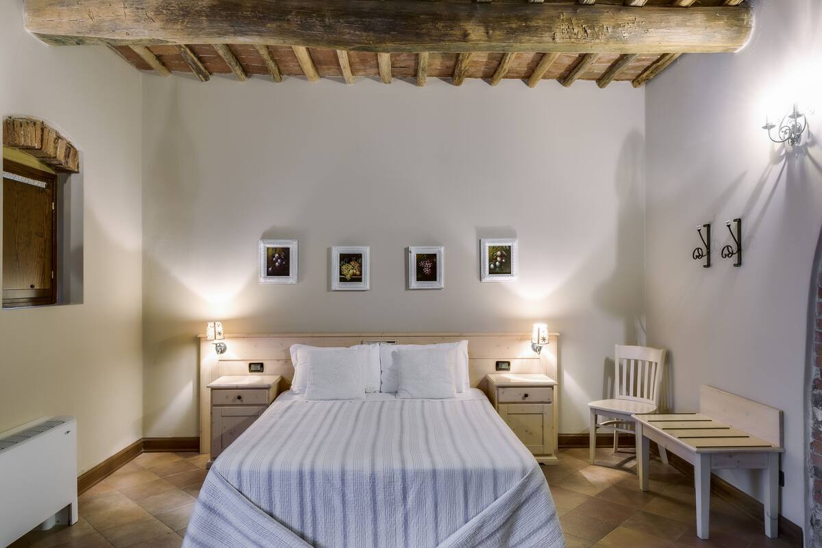 A bedroom with a wooden ceiling, a bed with white bedding, bedside tables, a chair, and wall-mounted picture frames.