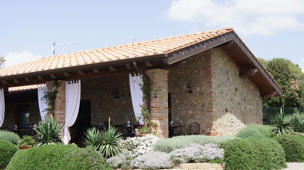 A rustic stone building with a tiled roof, a covered patio with outdoor seating, and well-maintained gardens.