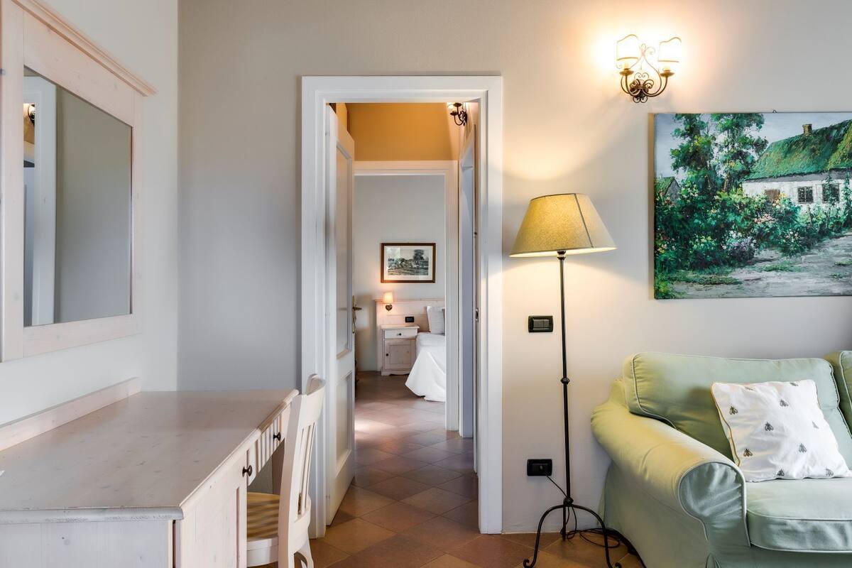 A hallway leading to a bedroom with a bed, a bedside table, a lamp, and a painting on the wall.