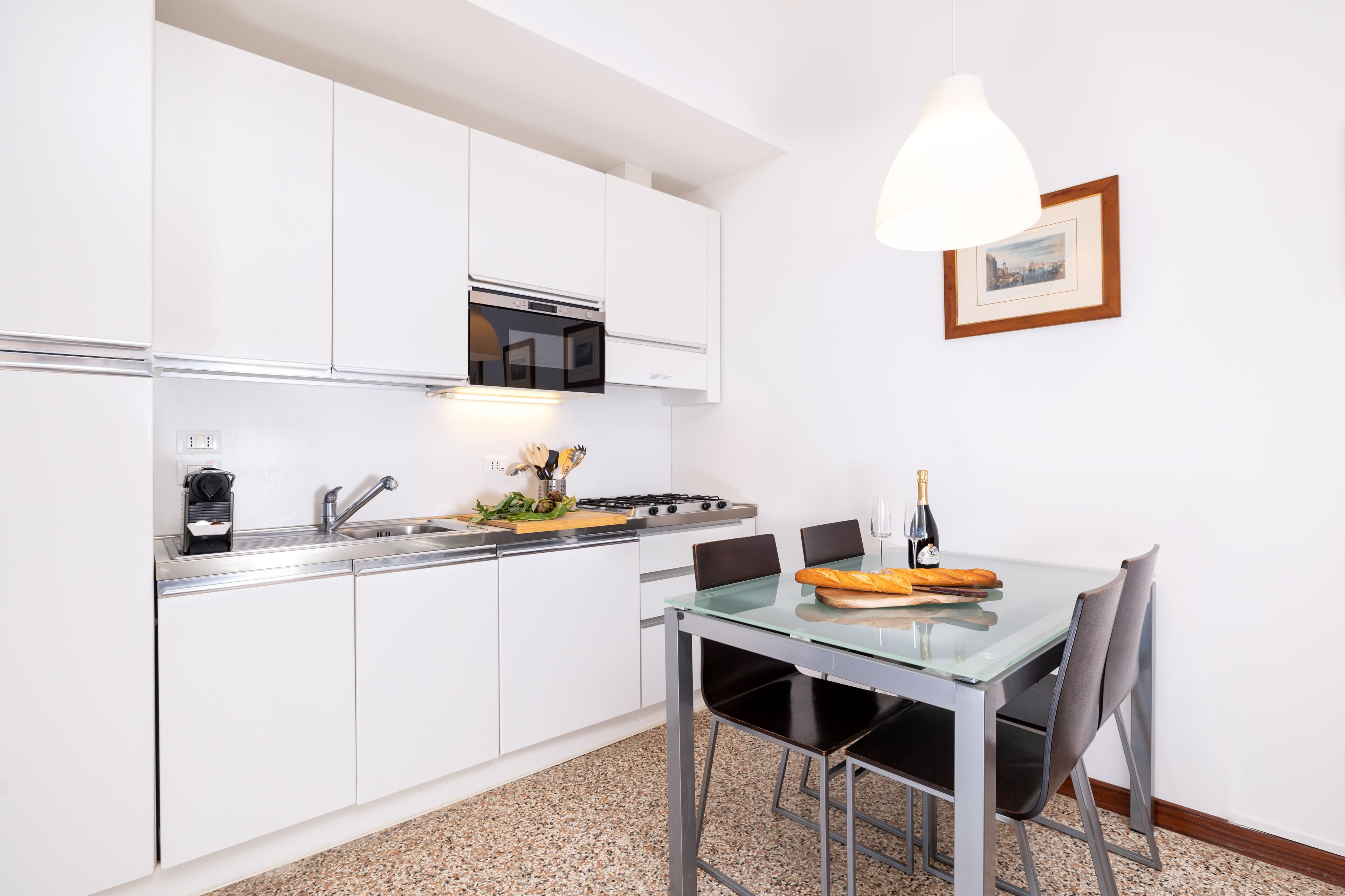 Une cuisine moderne avec des meubles blancs, un évier en acier inoxydable et une table à manger en verre, accompagnée de chaises.