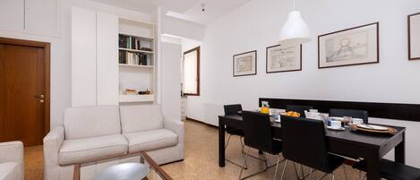 A modern living room with a sofa, a dining table with chairs, and a kitchen area with cabinets and a refrigerator.