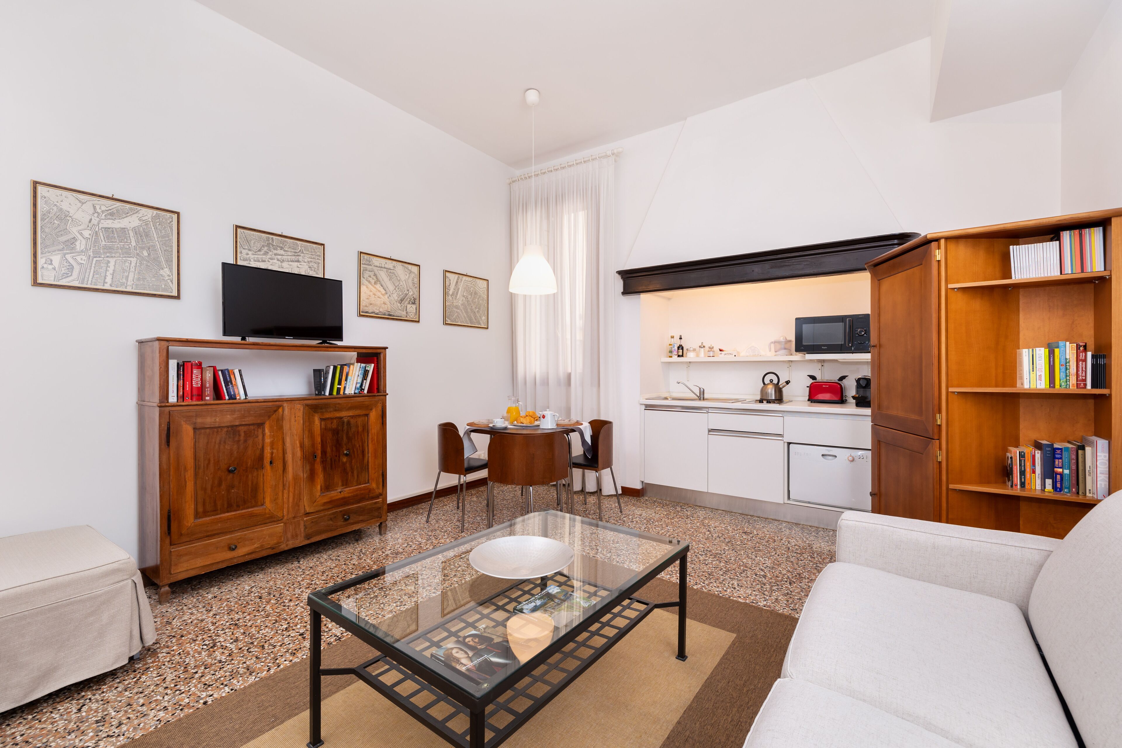 A modern living room with a glass coffee table, a sofa, a wooden bookshelf, a TV, and a kitchen area with a microwave and a kettle.