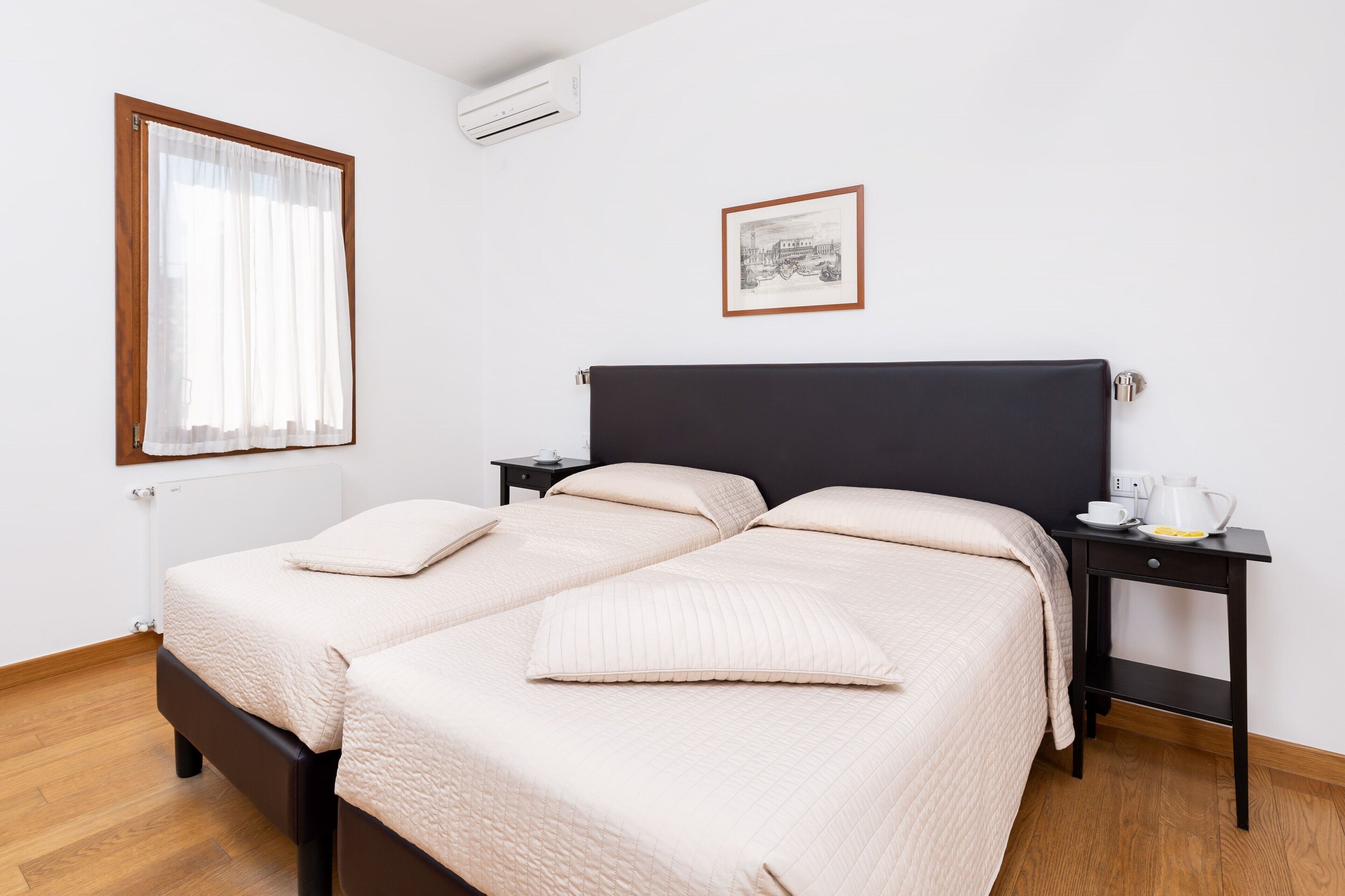 Een tweepersoonsbed met een donker hoofdeinde, twee nachtkastjes met een witte beker en een kleine lamp, een lijst met een afbeelding aan de muur, en een raam met transparante gordijnen.