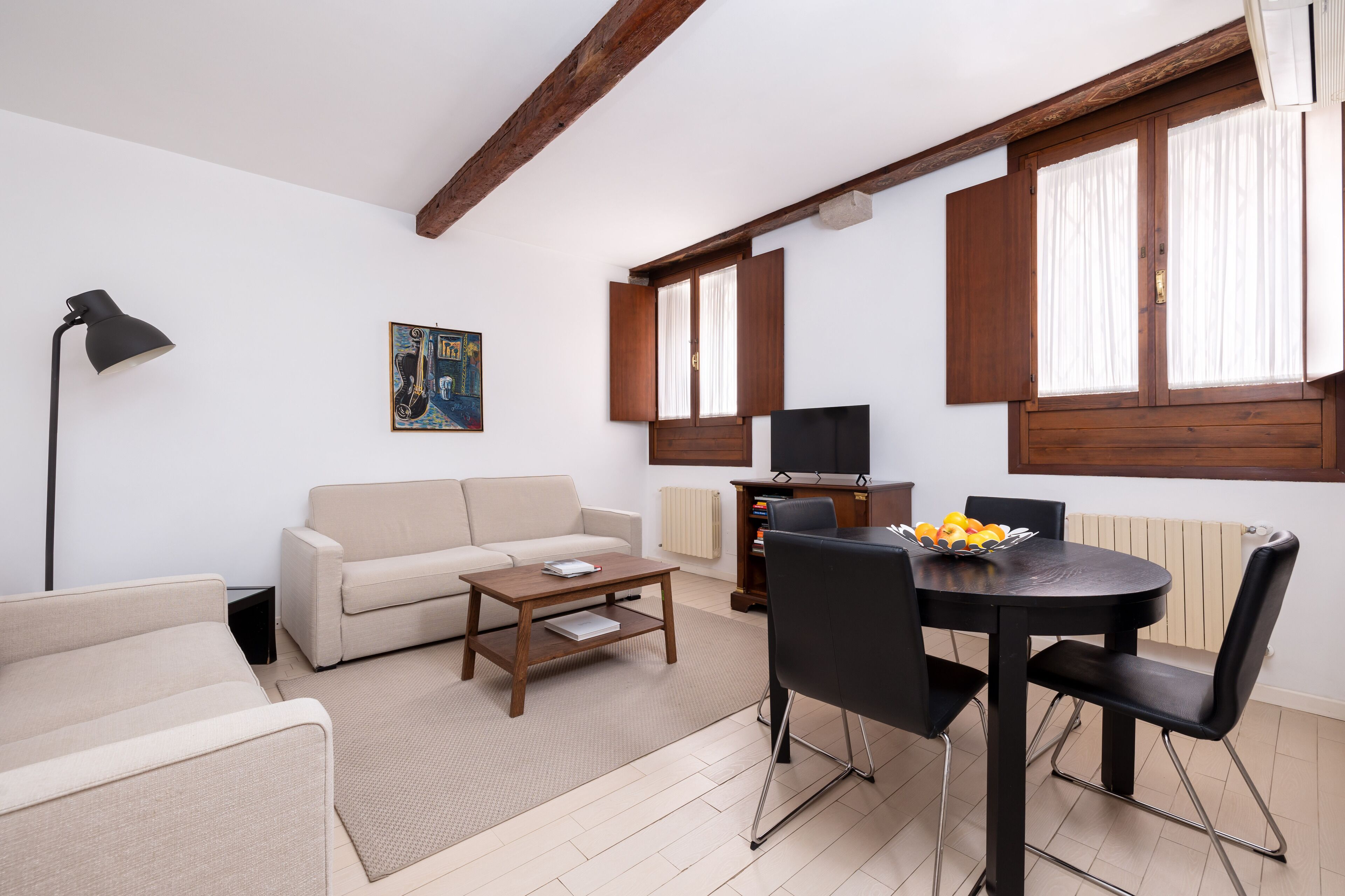 A living room with a sofa, coffee table, dining table, and chairs. There is a painting on the wall, a lamp, and a radiator.