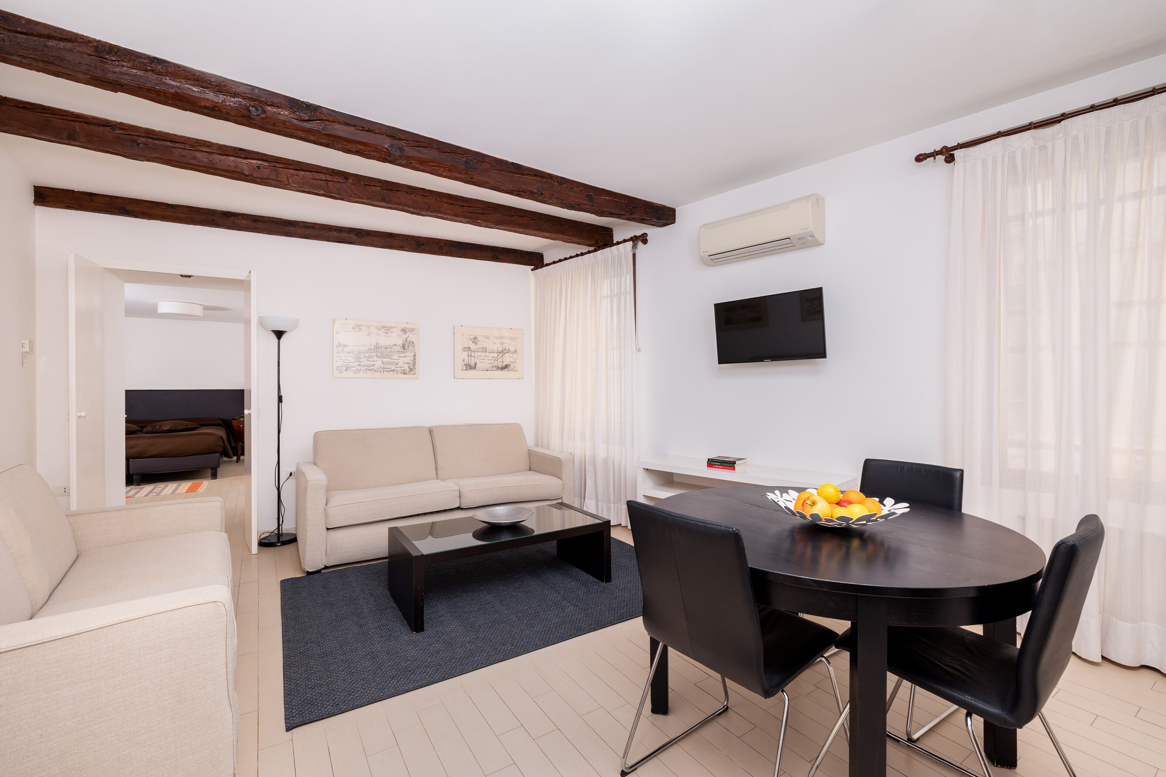 A modern living room with a sofa, coffee table, and dining table with chairs. There is a flat-screen TV mounted on the wall and a window with sheer curtains.