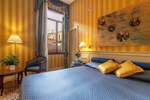 A hotel room with a large bed, two bedside lamps, a chair, a small table with a plant, and a window with curtains.