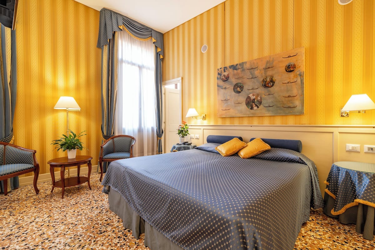 Ein Hotelzimmer mit einem großen Bett, zwei Sesseln, einem kleinen Tisch mit einer Pflanze und einem Bild an der Wand.
