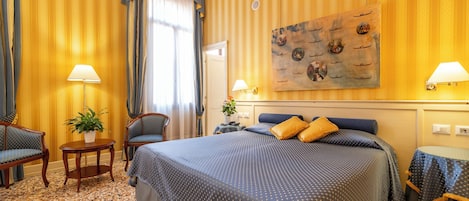 A hotel room with a large bed, two chairs, a small table with a plant, and a painting on the wall.
