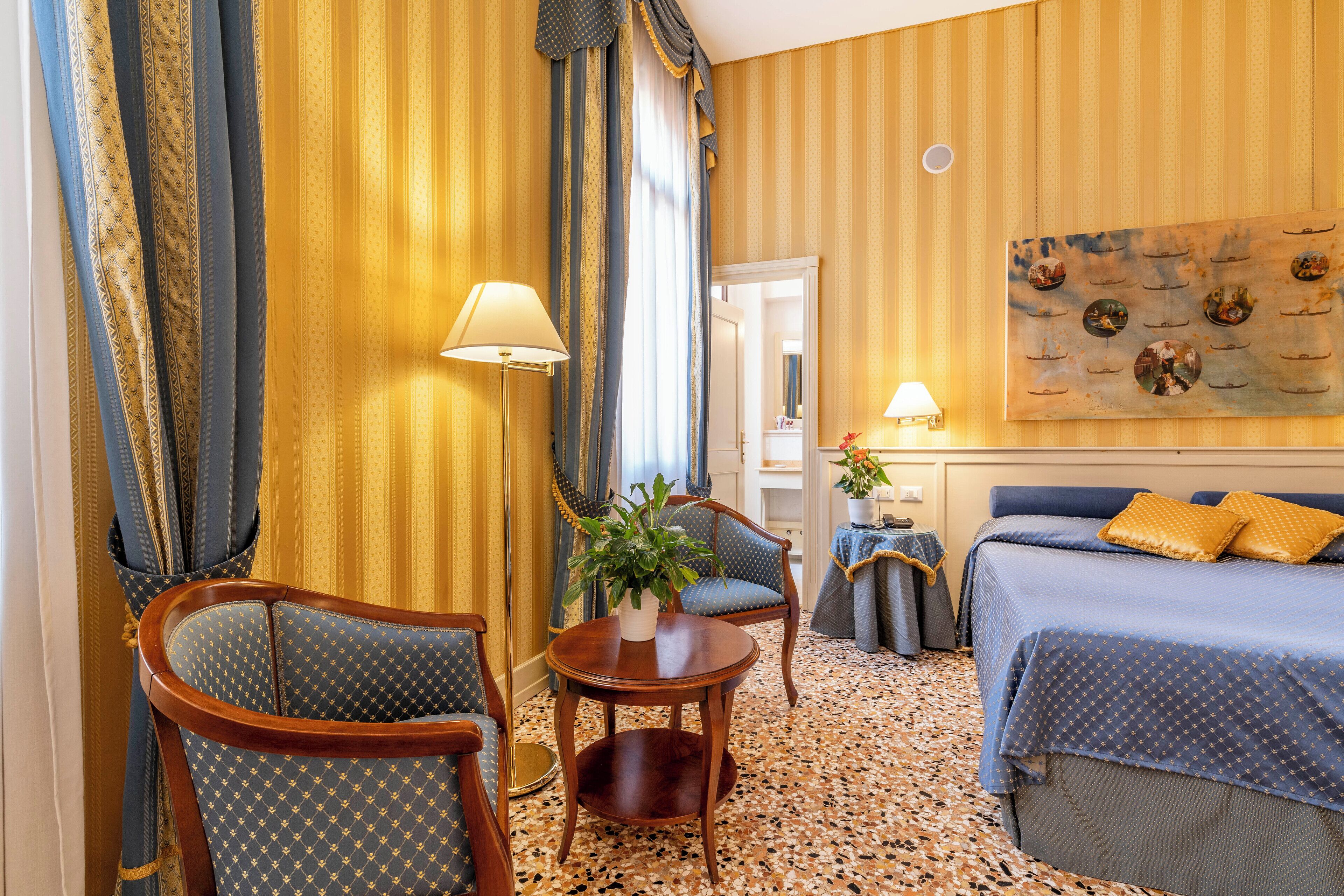 A hotel room with a bed, two armchairs, a small table with a plant, and a painting on the wall.