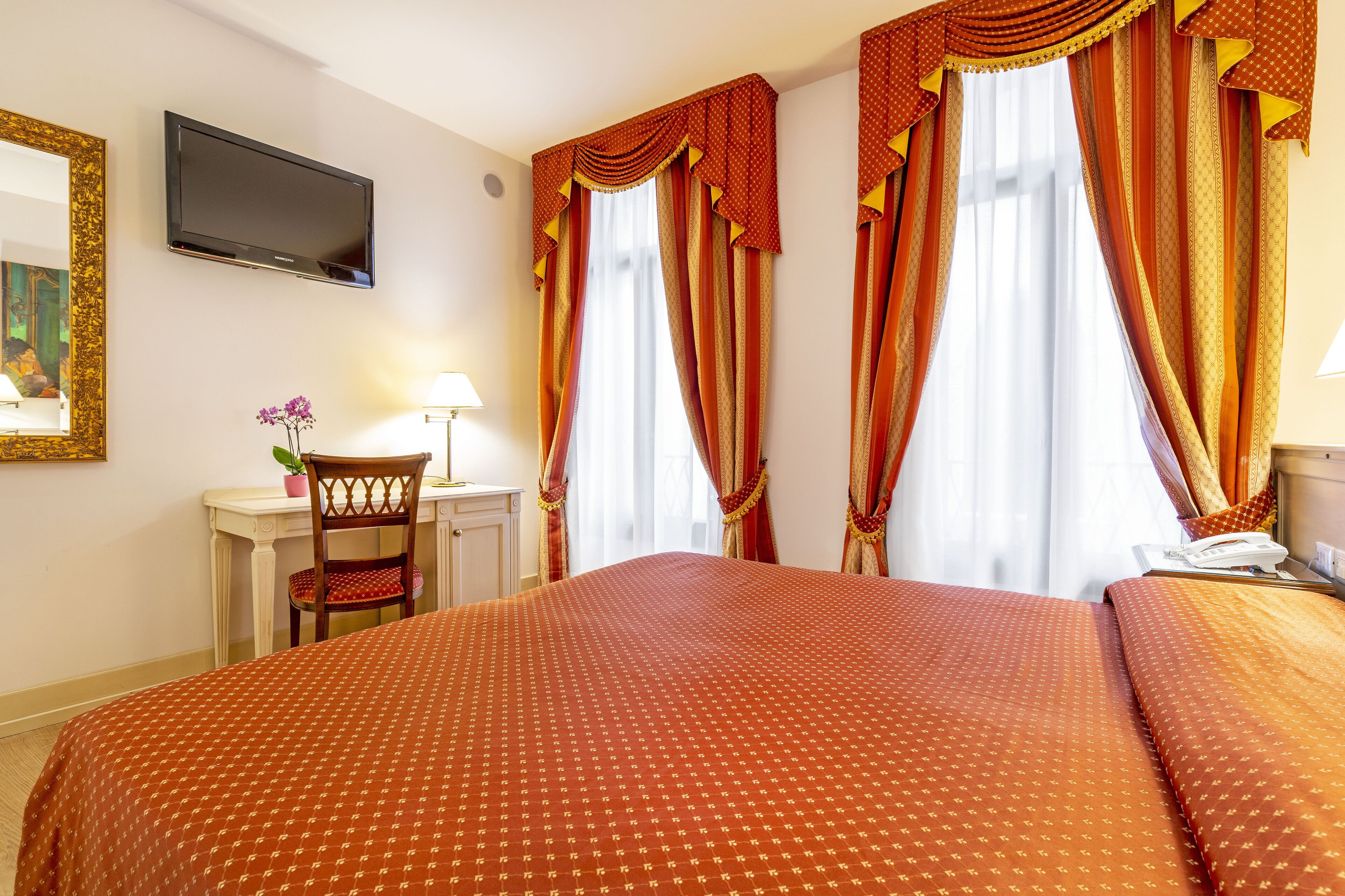 Quarto de hotel com uma cama grande, uma escrivaninha com cadeira, televisão fixada na parede e cortinas nas janelas.