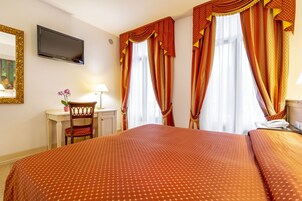 Quarto de hotel com uma cama grande, uma escrivaninha com cadeira, televisão fixada na parede e cortinas nas janelas.