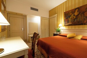 Quarto de hotel com cama, escrivaninha, cadeira, um quadro na parede e uma luminária.