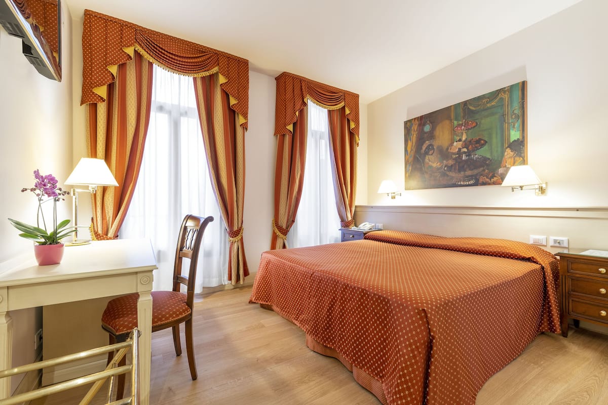 Ein Hotelzimmer mit Bett, Schreibtisch, Stuhl, Zimmerpflanze und einem Kunstwerk an der Wand.